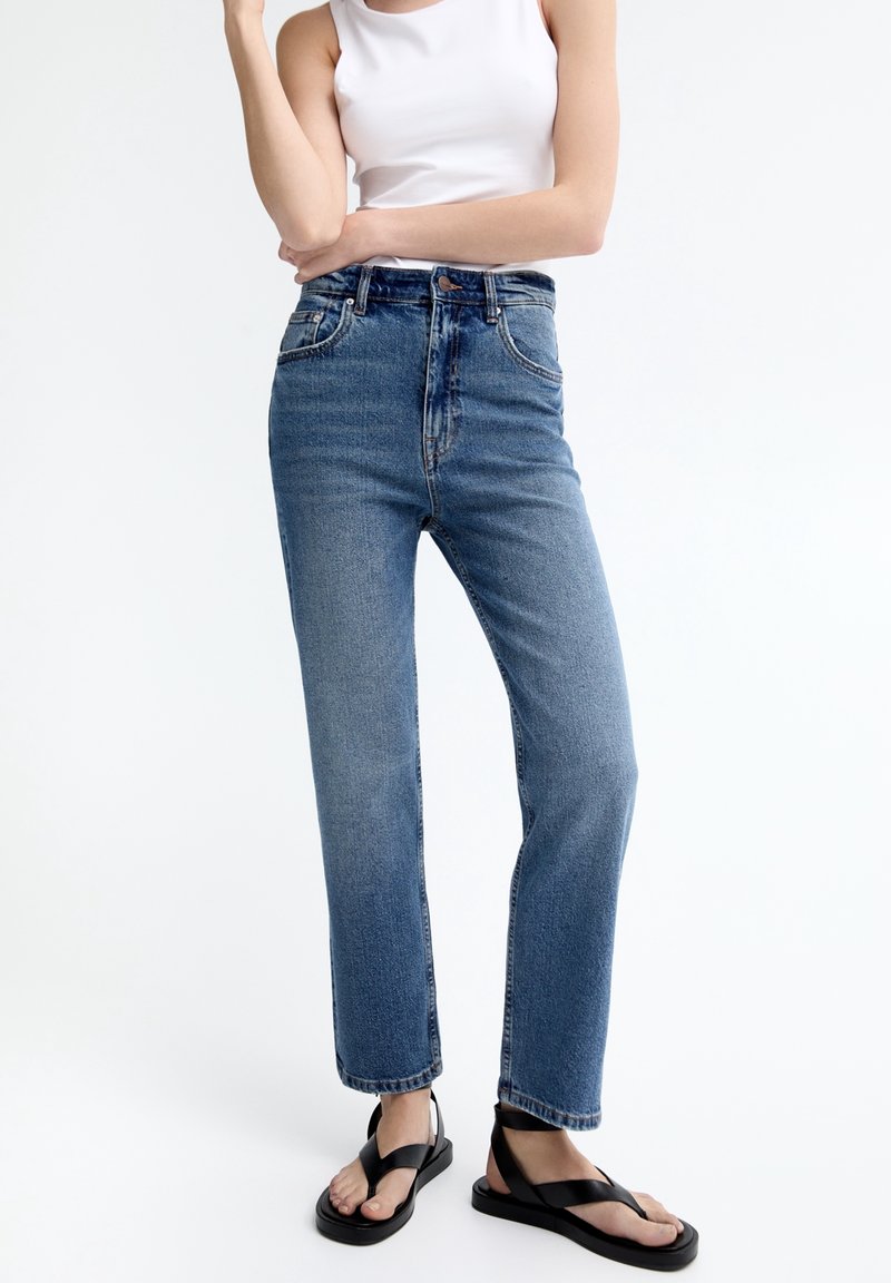 Femme portant un jean bleu taille haute, coupe droite, un débardeur blanc et des sandales plates noires, debout devant un fond blanc.