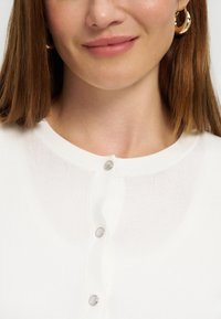 Γυναίκα με ίσια καστανά μαλλιά, φορά χρυσούς κρίκους στα αυτιά και λευκό πλεκτό πουλόβερ με στρογγυλά κουμπιά.