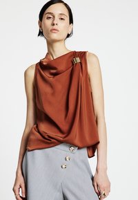 Top senza maniche in tessuto solido color ruggine con drappeggio e spilla decorativa dorata. Indossato con pantaloncini a vita alta grigio chiaro con bottoni decorativi.