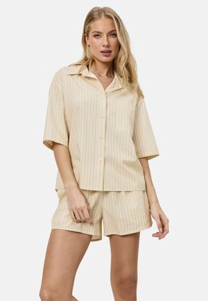 Blonde vrouw die een beige-wit gestreept shirt met korte mouwen en knopen draagt en bijpassende korte broek, poseert tegen een effen achtergrond.