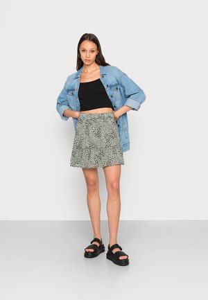 Jaqueta de ganga, top cropped preto ajustado, saia verde com pintas pretas e sandálias de plataforma pretas. Modelo está de pé com as mãos na cintura.