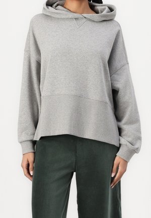 Žena v světle šedé mikině s kapucí s žebrovaným lemem a manžetami, v kombinaci s tmavě zelenými manšestrovými kalhotami, stojící na bílém pozadí.