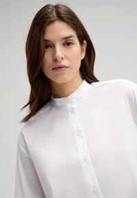 Weiße, knopflose Bluse mit Stehkragen, die eine glatte Textur und einen subtilen Glanz aufweist und für eine lockere Passform konzipiert ist.