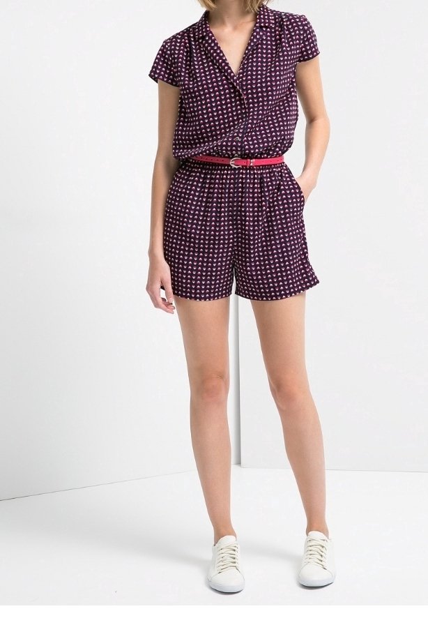 Jumpsuit de manga corta morado con un patrón de corazones blancos, cintura ceñida y un cinturón rosa. Combinado con zapatillas blancas, sobre un fondo liso.