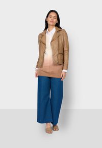 Braune Leder-Motorradjacke, weißes Hemd, Verlaufspullover in Beige, weite blaue Jeanshose und hellbraune Sandalen. Auffällige Schichtung und Texturen.