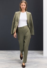 Olu zaļa pieguloša žakete un atbilstošas bikses kombinācijā ar baltu topu. Apģērbu papildina brūnas dzīvokļu kurpes uz neitrāla fona.