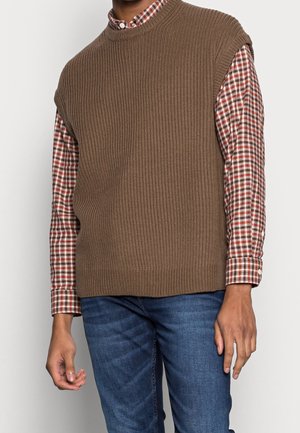 Un pull sans manches à côtes marron porté sur une chemise à carreaux rouges et blancs, associé à un jean bleu. Texture lisse avec des motifs contrastants.