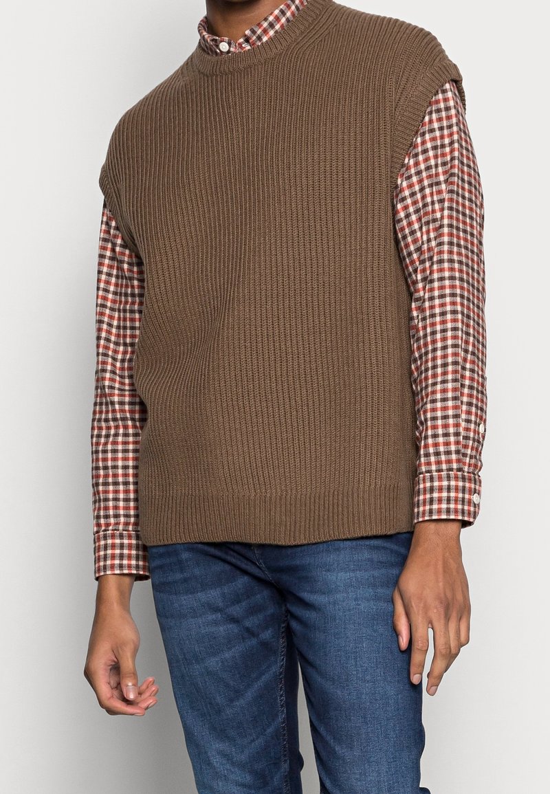 Un pull sans manches à côtes marron porté sur une chemise à carreaux rouges et blancs, associé à un jean bleu. Texture lisse avec des motifs contrastants.