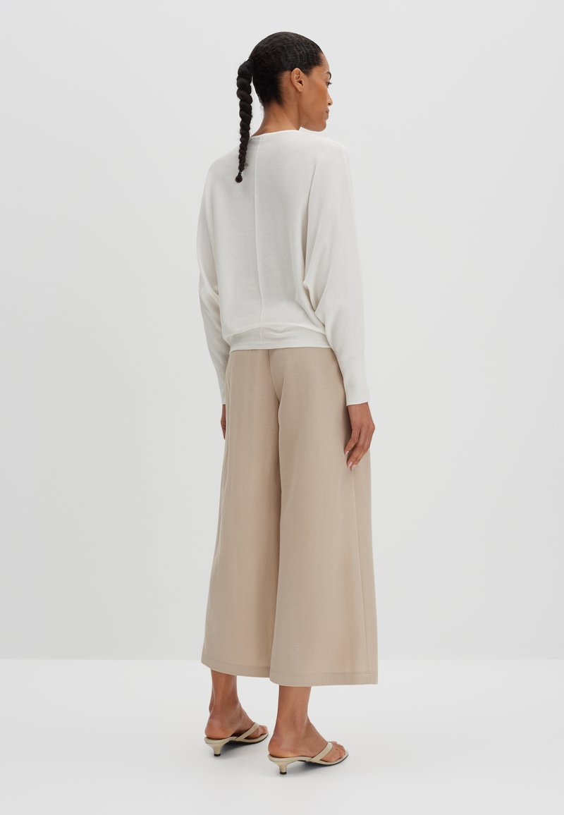 Biała, luźna bluzka z długim rękawem w połączeniu z beżowymi spodniami o szerokich nogawkach, charakteryzującymi się gładką fakturą i subtelnym marszczeniem.