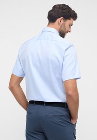 Camicetta azzurro chiaro a maniche corte con colletto classico, caratterizzata da una texture liscia. Abbinata a pantaloni blu scuro, che mostrano una vestibilità elegante.
