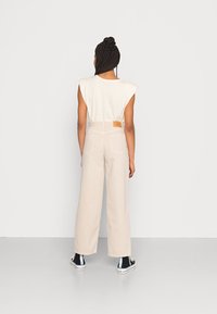 Calças largas beiges com uma cintura alta e uma pequena etiqueta de couro castanho nas costas. Combinadas com uma blusa larga sem mangas de cor creme.