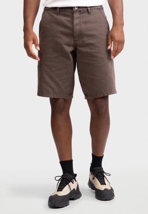 Άνδρας φορά καφέ σόρτς μέχρι το γόνατο, μαύρες κάλτσες και μπεζ-μαύρα αθλητικά, στέκεται σε απλό ανοιχτό φόντο.