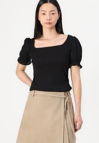 Top corto nero con scollo quadrato e maniche a sbuffo testurizzate, abbinato a una gonna avvolgente beige chiaro con dettaglio a fiocco.