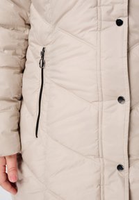 Beige gewatteerde jas met een ritssluitingzak, drukknopen en diagonale stikpatronen voor extra textuur en design.