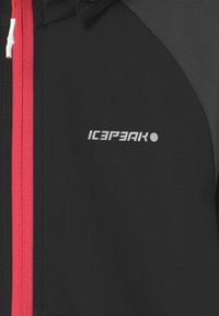 Sort atletisk jakke med rød lynlås og detaljer. Har en glat tekstur, ergonomisk design og et iøjnefaldende logo.