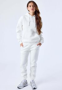 Sudadera blanca con cordones y bolsillo canguro combinada con pantalones deportivos blancos con puños elásticos, ambos en un tejido suave y texturizado.