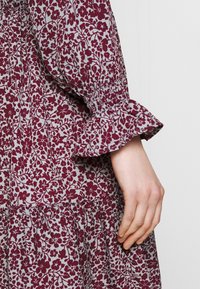 Långärmad klänning med ett vinrött blommönster på en ljusgrå tyg. Har samlade manschetter och en flerlagers nederdel.