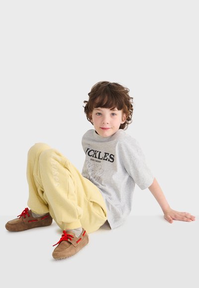 Kind mit lockigem braunem Haar sitzt auf dem Boden und trägt ein graues T-Shirt mit Aufdruck, gelbe Jogginghosen und braune Schuhe mit roten Schnürsenkeln.