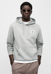 Γκρι φούτερ με κουκούλα, τσέπη μπροστά, κορδόνι στην κουκούλα και λογότυπο. Το μοντέλο φορά επίσ ης μπεζ γυαλιά ηλίου και καρό παντελόνι με μπλε λεπτομέρειες.