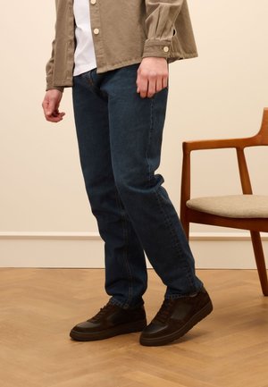 Dark blue denim jeans paired with a light-colored shirt and khaki jacket. Brown sneakers with textured accents, standing on wooden flooring.