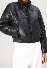 Czarna puchowa kurtka o błyszczącym wykończeniu, krótki fason, wysoki kołnierz i zapięcie na zamek. Posiada elastyczny dół oraz boczne kieszenie.