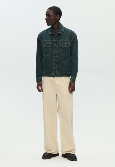 Uomo che indossa una giacca di jeans scura, pantaloni beige a gamba larga e scarpe nere, in piedi e guardando di lato contro uno sfondo bianco uniforme.