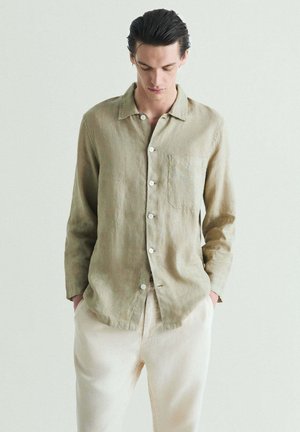 Hombre con cabello oscuro peinado hacia atrás, vistiendo una camisa beige abotonada y pantalones crema claros, con las manos en los bolsillos, mirando hacia abajo.