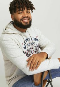 Hanorac gri deschis cu un buzunar frontal, având text albastre și roșii. Modelul este așezat pe un scaun din lemn, purtând blugi decolorati.