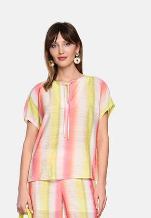 Vrouw die een pastelgestreepte top met korte mouwen en bijpassende broek draagt, met grote ronde gouden oorbellen, en een gele handtas vasthoudt.