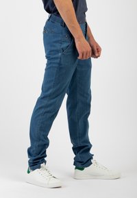 Pantaloni in denim blu con vestibilità slim, cintura elastica, tasche anteriori e scarpe da ginnastica bianche con dettagli verdi. Texture liscia e design contemporaneo.
