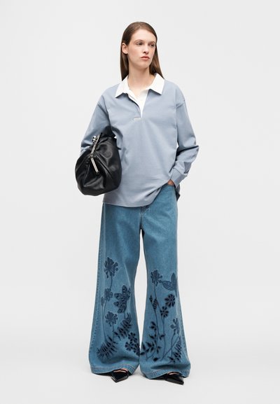 Ανοιχτό μπλε πουλόβερ με λευκό γιακά, σε συνδυασμό με τζιν παντελόνι φαρδύ με σκοτεινή ανθισμένη κεντήματα. Το μοντέλο κρατά μια μαύρη τσάντα.