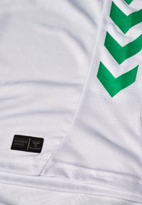 Tessuto di maglia sportiva bianca con strisce a chevron verdi sul lato e un'etichetta rettangolare nera della marca Hummel con la scritta "Authentic Apparel".