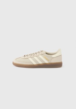 Sneaker beige Adidas con tre strisce bianche, suola in gomma color chewing gum e testo dorato "MÉTARIAL" sul lato, vista dal profilo sinistro.