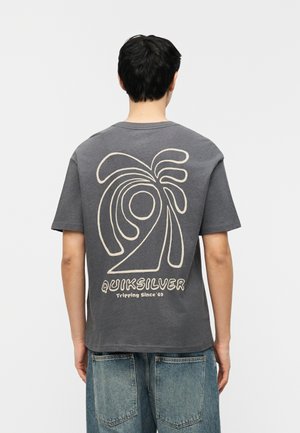 Mies, jolla on harmaa Quiksilver-t-paita, jossa on abstrakti aaltokuvio ja teksti "Tripping Since '69" takana, yhdistettynä sinisiin farkkuihin.