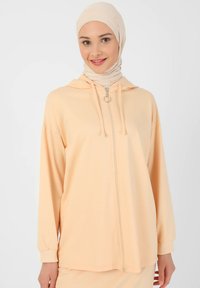 Light peach zip-up hoodie made of soft fabric, featuring a hood, long sleeves with ribbed cuffs, and a circular zipper pull detail.