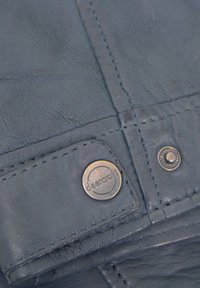 Nahaufnahme eines dunkelblauen Lederjackenarms mit genähten Nähten und einem metallischen Knopf, der mit "deercraft" graviert ist.