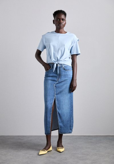 Chemisier bleu clair à manches courtes avec un nœud devant, associé à une jupe en denim de mi-longueur avec une fente avant, et des ballerines jaunes à bout pointu.