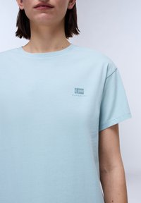 Persoon in een lichtblauw Napapijri t-shirt met een klein geborduurd logo op de borst tegen een effen achtergrond.