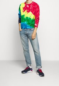 Flerfärgad tie-dye sweatshirt i röd, gul, grön och blå, tillsammans med ljusblå jeans och marinblå sneakers med röda accenter.
