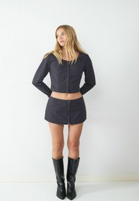 Schwarz gestreifter, verkürzter Blazer mit langen Ärmeln, kombiniert mit einem passenden Minirock. Beide haben eine glatte Textur, ergänzt durch kniehohe schwarze Stiefel.