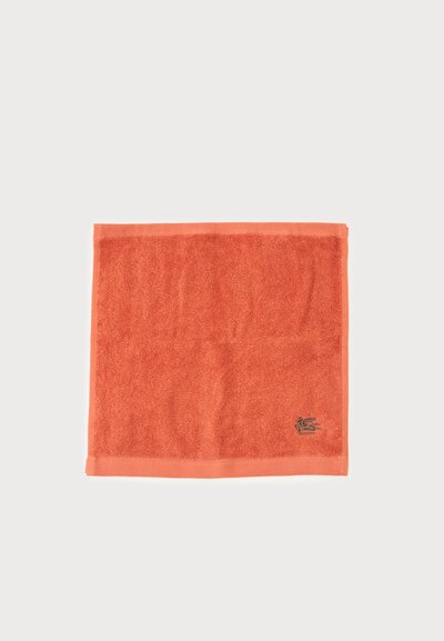 Carré de gant de toilette de couleur corail avec un petit logo brodé dans un coin sur un fond clair uni.