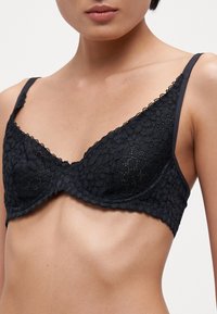 Melnas mežģīņu bralette ar trīsstūra formas kausiem, viļņotām malām, plānām siksnām un ziedu rakstu visā audumā.