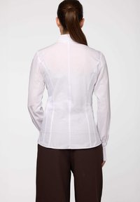Blusa bianca su misura con maniche lunghe, dotata di colletto alto e cuciture verticali per una silhouette sagomata. Tessuto leggero e morbido; vista posteriore.