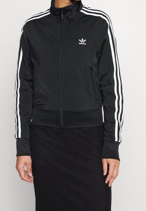 Sort Adidas træningsjakke med hvide striber på ærmerne, lynlås foran og logo på venstre bryst, båret af en person stående foran en ensfarvet baggrund.