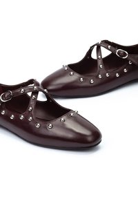 Zapatos de piel borgoña con punteras puntiagudas, presentan un diseño con recortes, correas cruzadas y tachuelas plateadas dispuestas decorativamente en la parte superior.