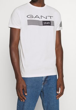 Man wearing white short-sleeve GANT T-shirt with black stripes and "1949" text, paired with blue jeans, against a plain background.
