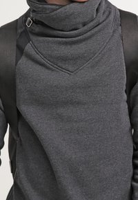 Gros plan d'une personne portant un pull gris foncé à col montant avec un détail de boucle et des bretelles de sac à dos noires sur les épaules.