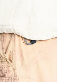Beige pants with front pocket, stitched details, and a black cord toggle attached to a white fabric hem above the waist area.