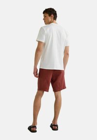 T-shirt blanc en coton à manches courtes, associé à un short en lin couleur rouille. Des sandales complètent la tenue, vues de derrière.