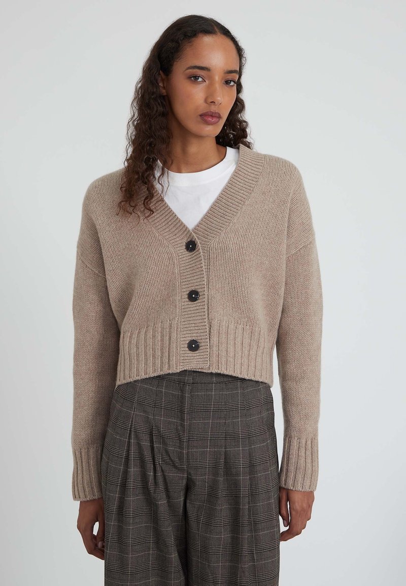 Beiges kurzes Strickjacke mit V-Ausschnitt, vier schwarzen Knöpfen und gerippten Bündchen. Kombiniert mit karierten Hosen. Glatte Textur.